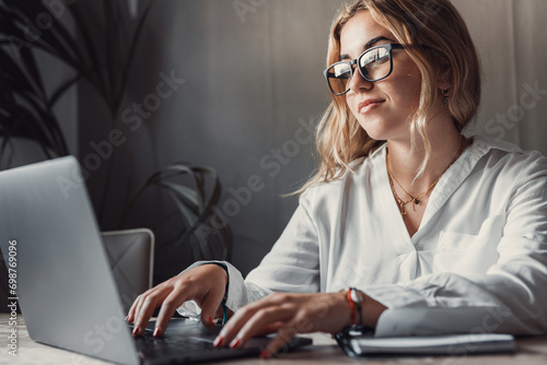 Happy young blonde business woman entrepreneur using computer looking at screen working in internet sit at office desk, smiling millennial female professional employee typing email on laptop at work photo
