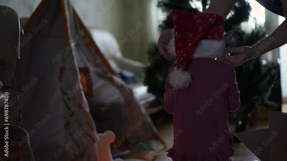 Christmas tree decoration, mother carefully puts a shiny red Santa Claus hat on her daughter, they go to the tree to hang toys and garlands, a children's wigwam stands nearby Christmas tree decoration, mother carefully puts a shiny red Santa Claus hat on her daughter, they go to the tree to hang toys and garlands, a children's wigwam stands nearby