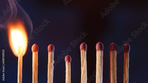 A row of matches with red gray on a dark studio background. The first match burns embraced by a flame. A tongue of flame burns the match, but the others remain intact. Macro image