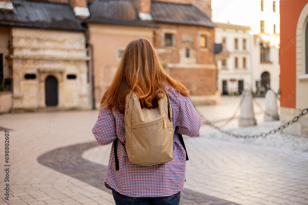 Naklejka premium Attractive young female tourist with backpack is exploring new city. Happy 30s woman walking on old city street. Traveling Europe in summer, urban background, rear view. Cracow, Poland