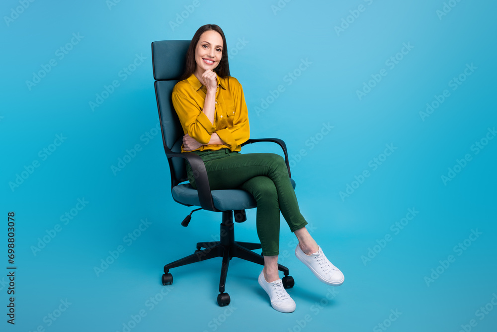 Full length photo of positive smart lady wear yellow shirt sitting office chair arm chin empty space isolated blue color background Full length photo of positive smart lady wear yellow shirt sitting office chair arm chin empty space isolated blue color background