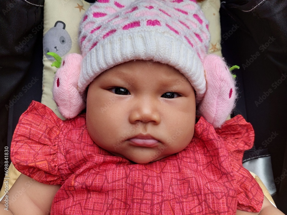 Cute Asian baby wearing a red dress and wearing a knitted hat, riding in a black stroller Cute Asian baby wearing a red dress and wearing a knitted hat, riding in a black stroller