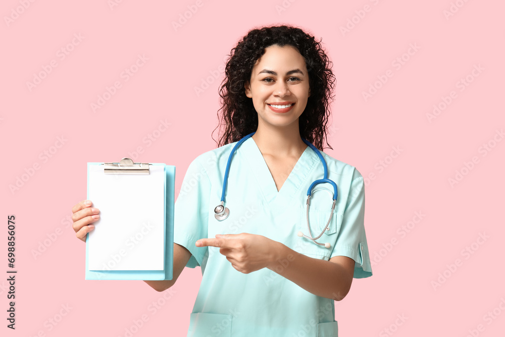 Beautiful young happy African-American female doctor with stethoscope pointing at clipboard on pink background Beautiful young happy African-American female doctor with stethoscope pointing at clipboard on pink background