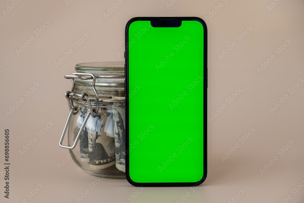 Obraz premium Vertical Green screen on modern mobile phone in background of glass jar full of American currency dollar banknotes on beige background. Cope space for text. Advertisement for application website