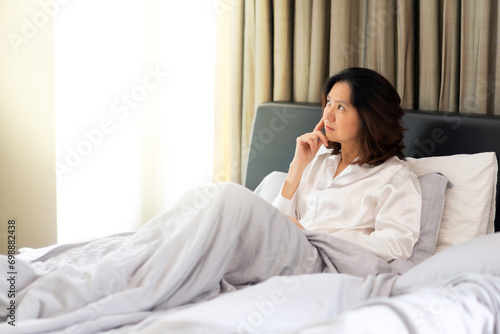 Asian middle edge women in white sleeping suit with clean skin, natural make-up, short black hair sitting on bed in bedroom pointing finger up and touch her face while thinking about future in morning