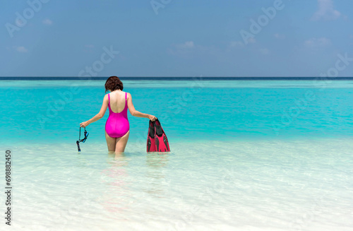 Back of Asian woman in pink swimsuit standing with mask and fins enjoy looking view of beautiful sea on hot summer day. Beautiful fit body girl in swimsuit relax on travel. Summer vacation concept.