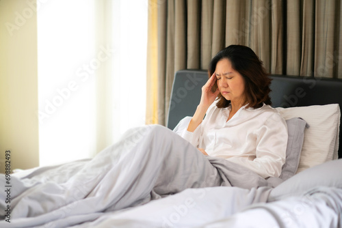 Portrait of stress middle aged woman got headache, close eyes and massage her head sitting on bed in bedroom. Asian Menopause women have painful Migraine and touch her temples, midlife crisis concept.
