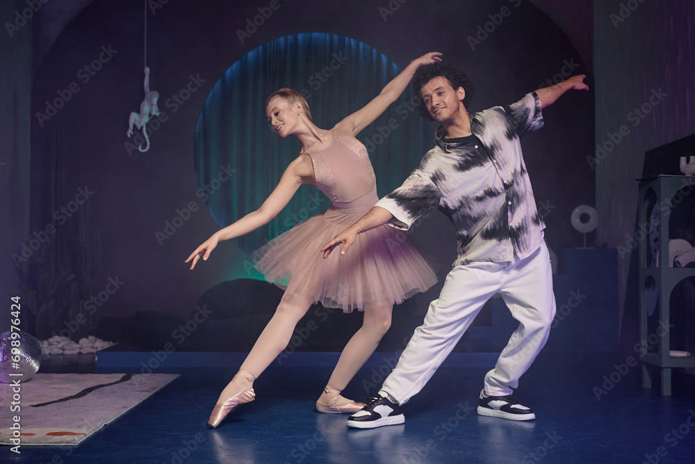 Wide shot of ballerina demonstrating ballet figure, young stylish man repeating after her