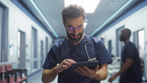 Tracking Shot of Latin Doctor Walking in Hospital Corridor, Using a Digital Tablet. Smiling Male Surgeon Checking Brain MRI Images Before Surgery, Revisiting his Notes, Greeting his Colleagues photo