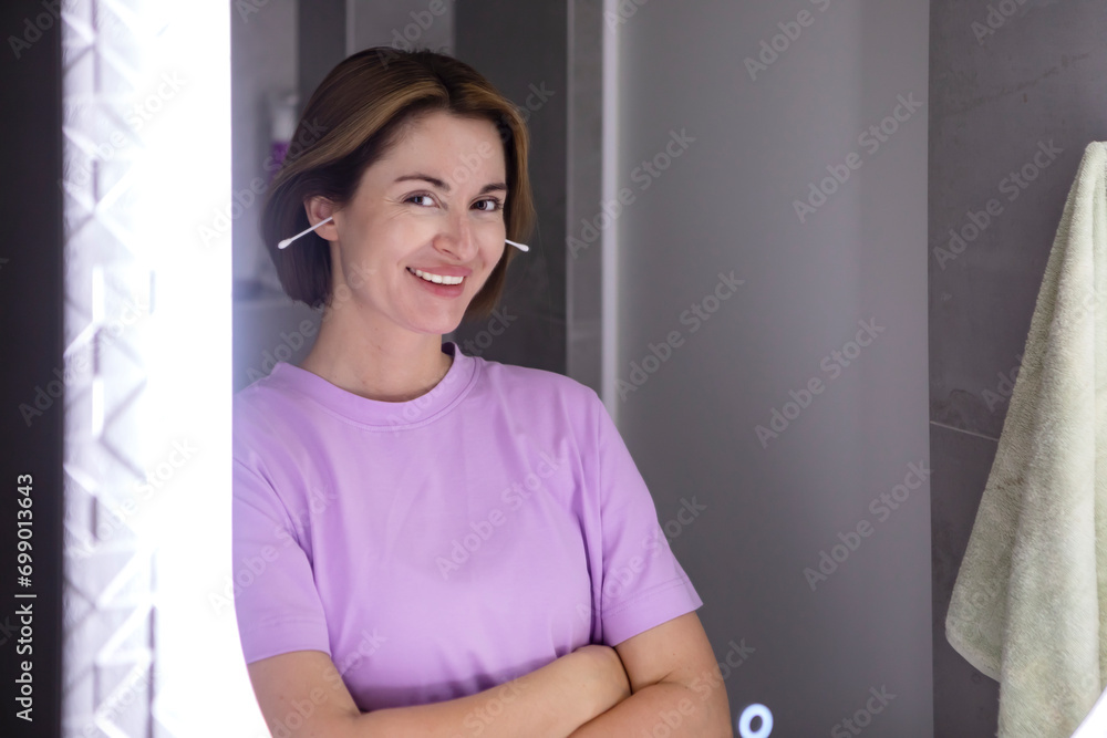 In the bathroom, a girl stands before the mirror, delicately using ear sticks, her focused reflection capturing a moment of personal care In the bathroom, a girl stands before the mirror, delicately using ear sticks, her focused reflection capturing a moment of personal care