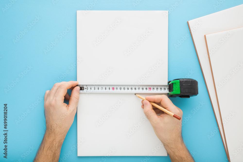 Man hand holding measure tape and measuring new white ceramic tile on blue table background. Closeup. Pastel color. Preparing for tiles laying. Point of view shot. Renovation process. Top down view. - obrazy, fototapety, plakaty 