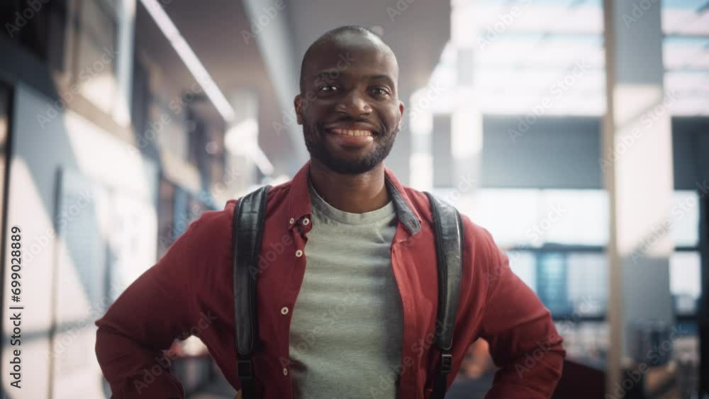 Portrait of a Cheerful Male Student Standing in a Modern Public Library. Young Black Man Looking at Camera and Smiling. Scholar Carrying a Backbag and Posing in University. Slow Motion Portrait of a Cheerful Male Student Standing in a Modern Public Library. Young Black Man Looking at Camera and Smiling. Scholar Carrying a Backbag and Posing in University. Slow Motion