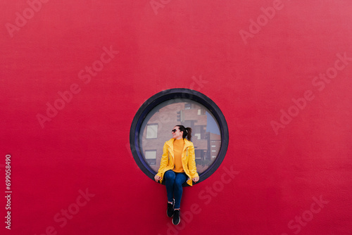 Woman in yellow rain coat sitting in porthole in red wall photo