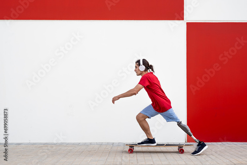 Man wearing wireless headphones and skateboarding with prosthetic leg in front of wall photo