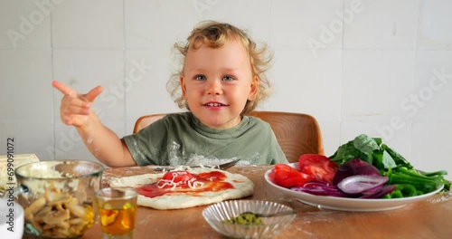 Young child making pizza playfully sprinkles grated cheese on dough, then curiously samples ingredient with hand-to-mouth taste test, gazing around with naive expression. Funny scene at kitchen photo