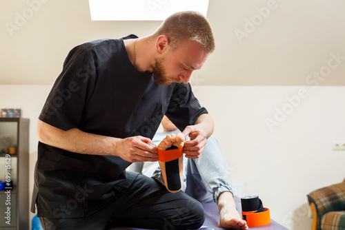 Male physiotherapist doctor massages the feet of a relaxed man sitting on a stretcher. He uses kinesio tape. Taping the foot © Дмитрий Ткачук