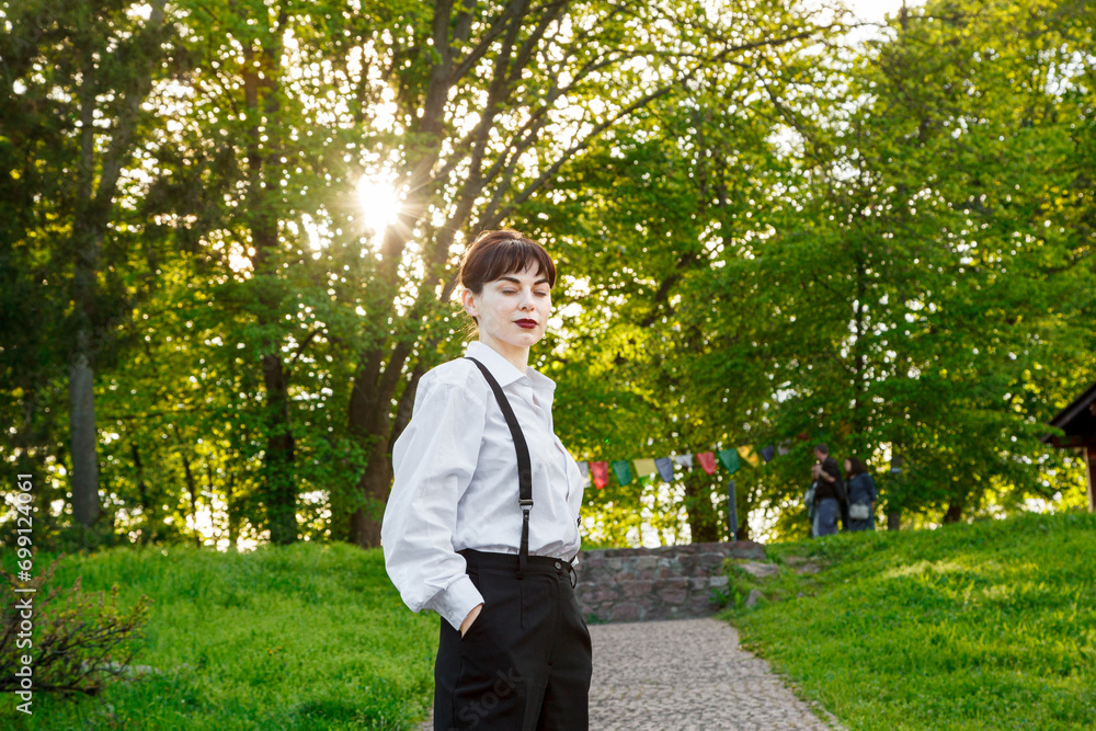 A beautiful girl in a white shirt, in black trousers with suspenders against the background of the sky and green grass A beautiful girl in a white shirt, in black trousers with suspenders against the background of the sky and green grass