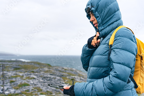 Latin woman sheltering from the wind with a hood while looking at the compass in a landscape with the sea in the background on the Irish coast