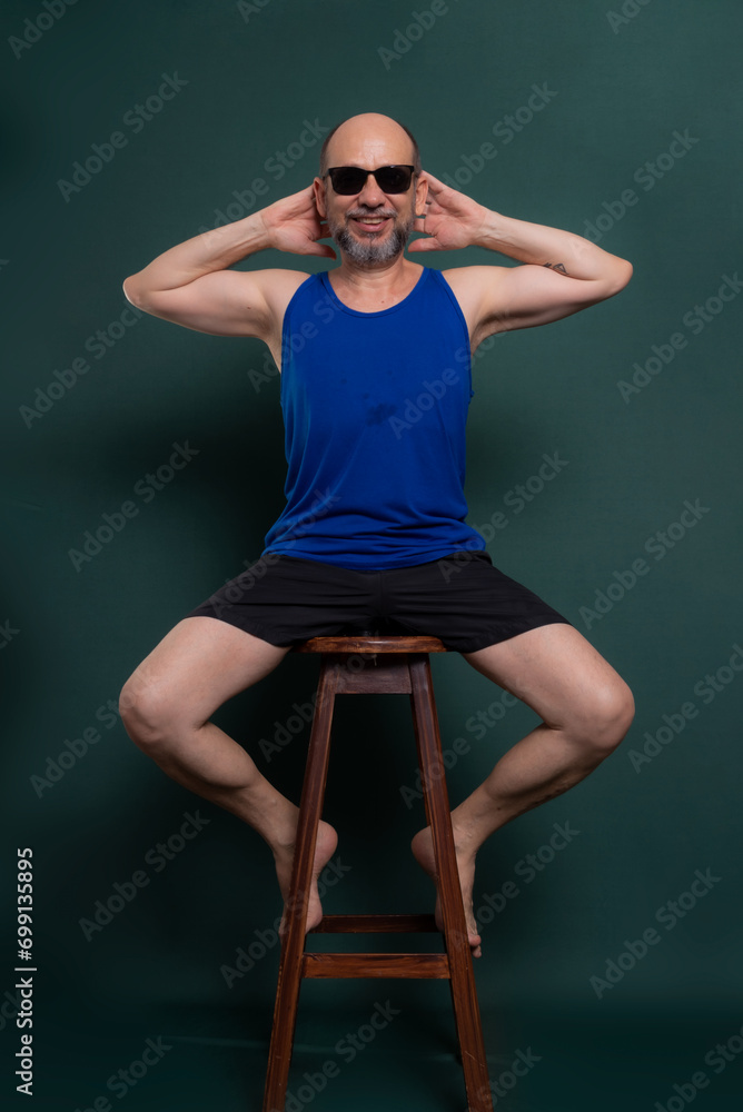 Bearded, bald white man wearing sunglasses sitting on wooden stool posing for photo.