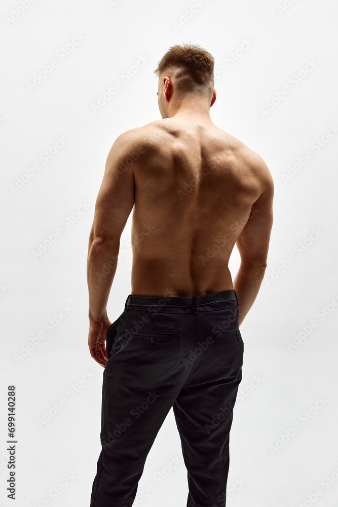 Rear view of shirtless male model with relief strong back, muscular body standing in pants isolated over white studio background. Concept of male beauty, body care, fitness, sport, health