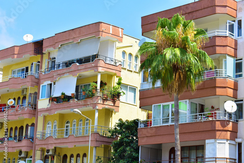 Fototapeta Naklejka Na Ścianę i Meble -  Modern residential house with red and yellow walls, balconies, plants and palm tree against a blue sky background. Sunny summer day. Citylife. Human life. Alanya, Turkey, July 2023
