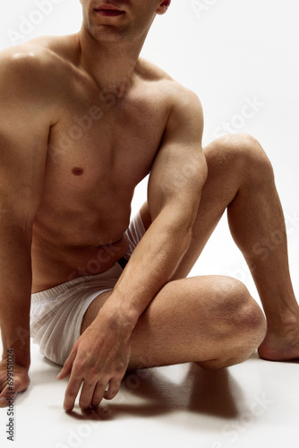 Cropped image of muscularly young shirtless man with relief body sitting in underwear, boxers isolated over white studio background. Concept of male beauty, body care, fitness, sport, health