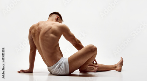 Rear view of young muscular man with relief body, strong back sitting shirtless in boxers isolated over white studio background. Concept of male beauty, body care, fitness, sport, health photo