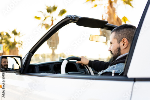 An adult man with a beard is driving a white antique convertible car at sunset. Cabriolet-style cars. Old white cabriot. #699186813