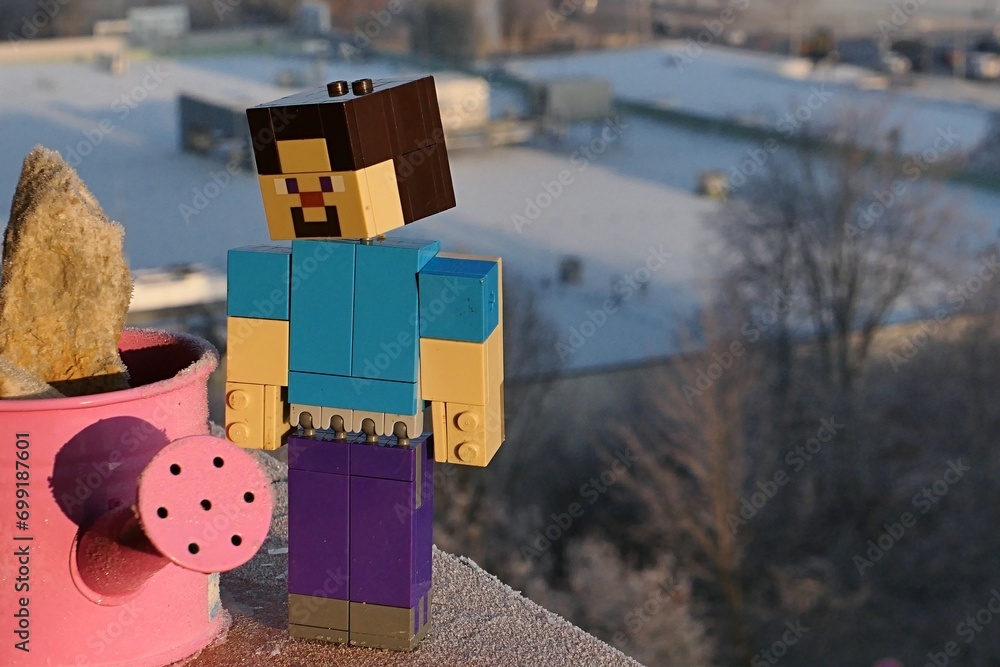 Naklejka premium LEGO Minecraft large figure of smiling Steve adoring frosted pink watering can, while standing on balcony cornice, winter building in background. Morning sunshine.