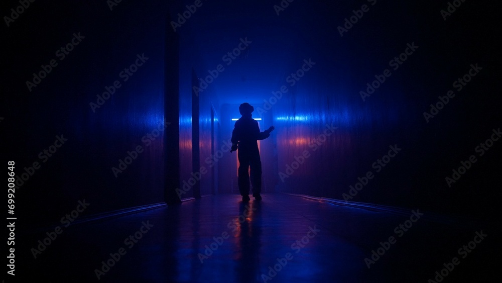Portrait of silhouette in the hallway neon light. Person in chemical protection suit walking cautiously in the dark corridor, holding up to wall