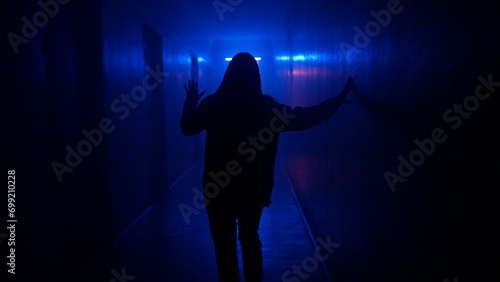 Portrait of female in hallway blue neon light. Close back shot woman walking cautiously in the dark corridor, holding up to wall.