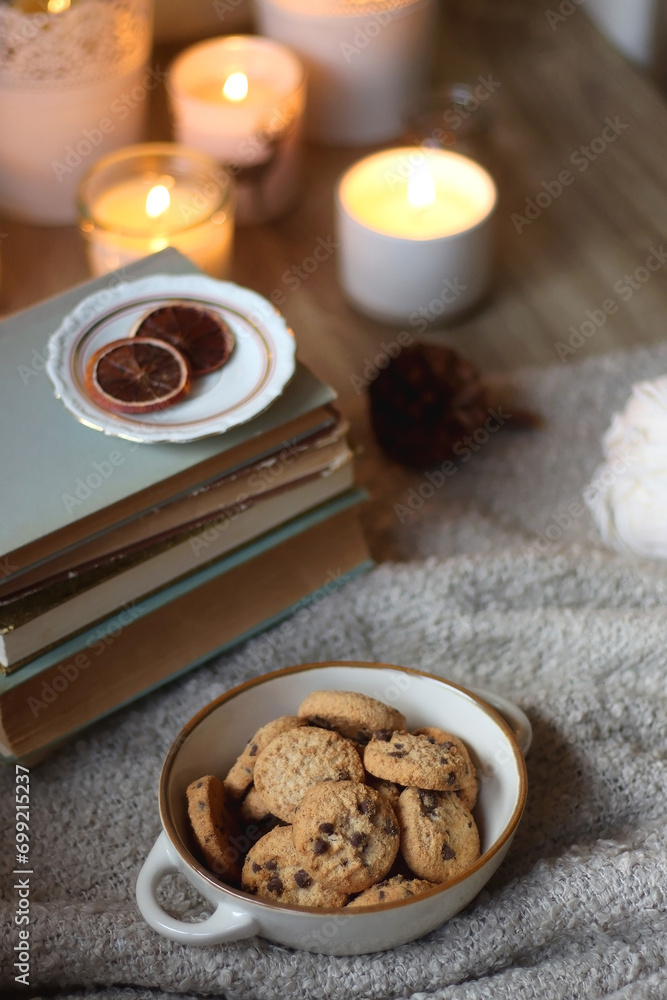 Bowl of cookies, cup of tea or coffee, plate of blueberries, dry oranges, wool yarn, pine cones, books, reading glasses, tablet, pen, soft blanket and lit candles on the table. Winter hygge at home. Bowl of cookies, cup of tea or coffee, plate of blueberries, dry oranges, wool yarn, pine cones, books, reading glasses, tablet, pen, soft blanket and lit candles on the table. Winter hygge at home.
