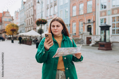 Redhead woman with paper map and phone on urban street. Young worried female traveler lost in the city using map and smartphone. Vacation concept by exploring interesting places to travel. © mdyn