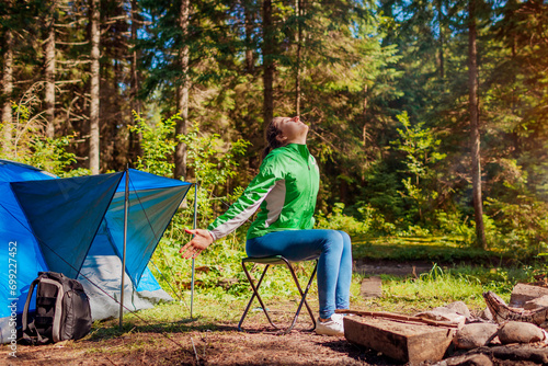 Happy woman relaxing by campfire in forest sitting next to tent with arms opened. Summer camping. Traveling alone