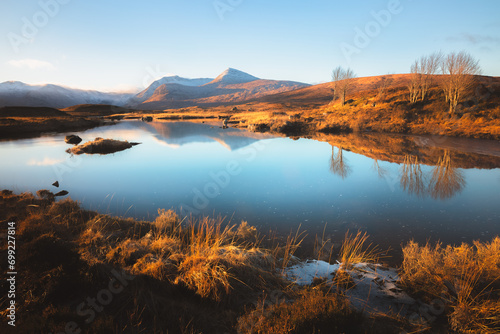 Gold light cast across a calm, tarn reflection and moorland at Rannoch Moor in the mountain landscape of Glencoe in the Scottish Highlands, Scotland, UK.