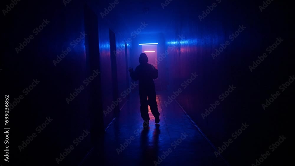 Portrait of silhouette in the hallway neon light. Person in chemical protection suit walks cautiously in the dark corridor, looking around facing camera. Portrait of silhouette in the hallway neon light. Person in chemical protection suit walks cautiously in the dark corridor, looking around facing camera.