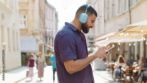 Happy relaxed overjoyed indian man in wireless headphones choosing, listening favorite energetic disco music in smartphone dancing outdoors. Guy tourist walking passes by urban sunshine city street photo