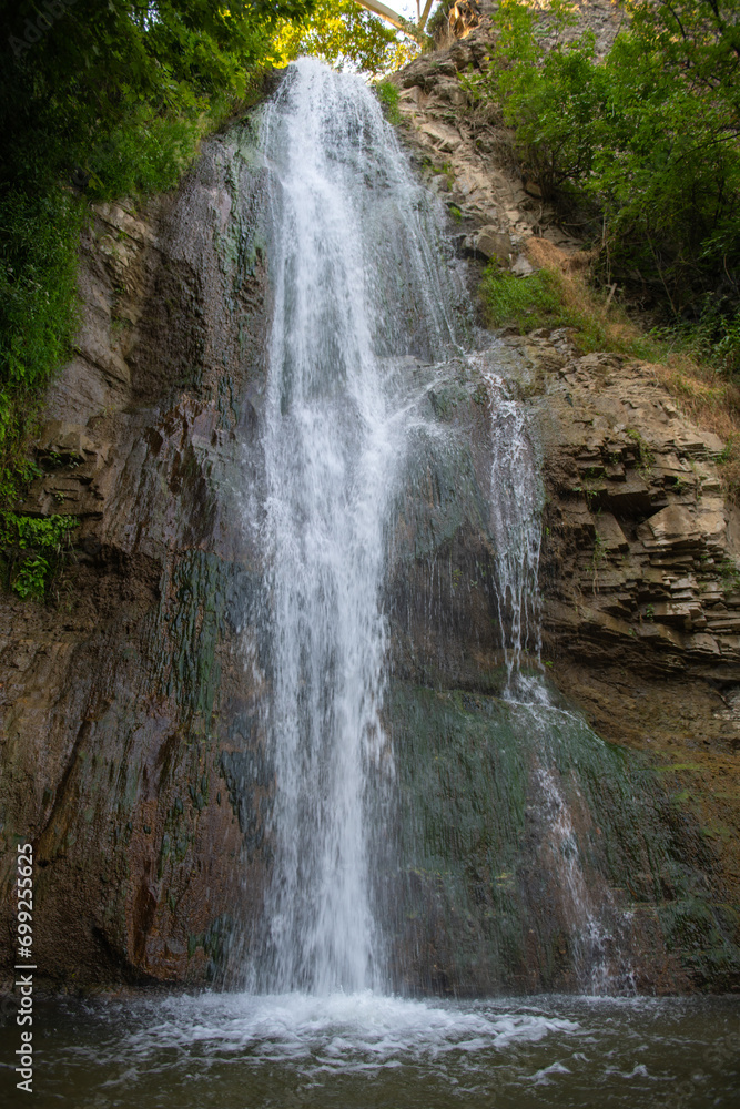 Against the background of green trees, the water of the waterfall flows Against the background of green trees, the water of the waterfall flows