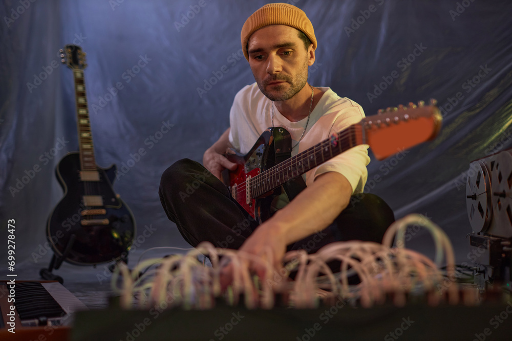 Obraz premium Medium full shot of male musician in yellow beanie sitting on studio floor adjusting musical equipment for electric guitar recording