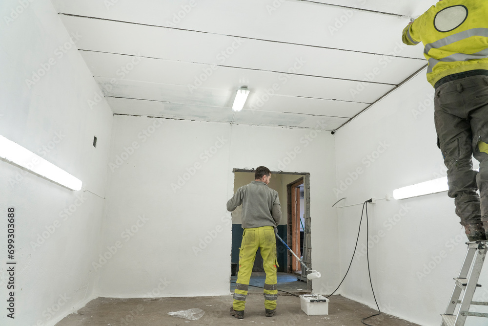 Two Workers in Protective Attire Paint a Spacious Room in Clean White Finish.