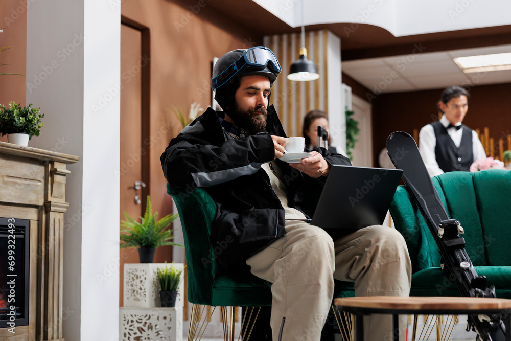 In luxury lounge area, man dressed in winter jacket enjoys coffee while using laptop to explore winter activities. Combination of technology and comfort adds excitement for tourist with skiing gear.
