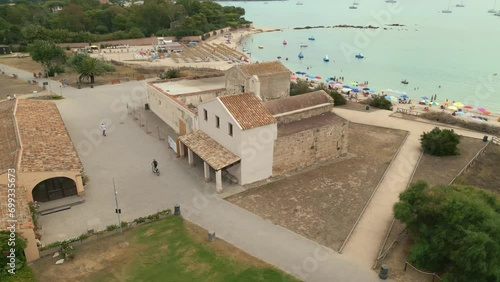 Aerial orbit around romanesque Church of Sant'Efisio (Chiesa di Sant'Efisio) in Nora, Pula, Sardinia, Italy. Beautiful beach behind, with colorful umbrellas, boats and people swimming. photo