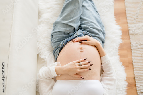 Cropped color photo taken from above of pregnant woman in unbuttoned blue jeans laying on white couch with hands on her bare pregnant belly aesthetic photo