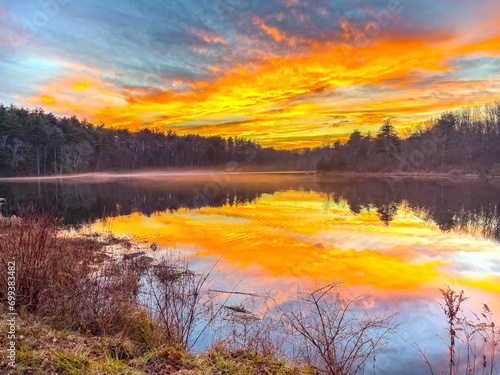Fototapeta Naklejka Na Ścianę i Meble -  A bright and colorful sunset with orange sky and clouds and a perfect reflection over the New England Pond. Lake shore with bushes in front of peaceful sun set. 