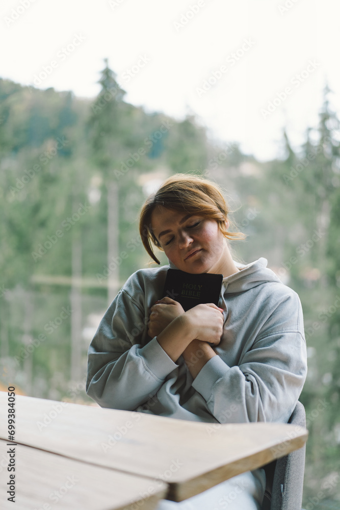 Fototapeta premium A girl reads the Bible against the background of a beautiful forest panorama. Christian woman holds holy bible in hands. Concept for faith, spirituality and religion.