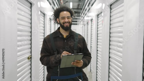Bearded smiling dark-skinned man, self storage worker in checkered T-shirt, work overall holding tablet in a typical climate controlled storage facility. High quality 4k footage photo