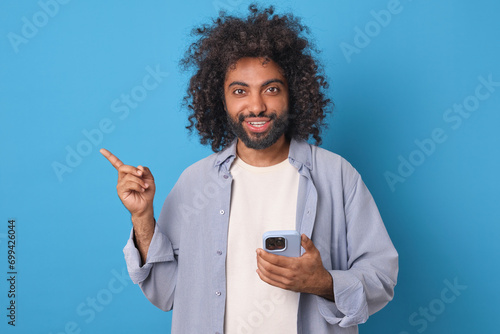 Young happy handsome Arabian man with mobile phone in hands smiles sincerely and points finger to side offering to download online store application for smartphone users stands in blue studio. photo