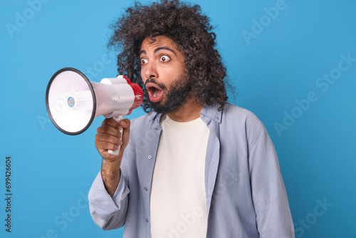 Young surprised curly Arabian man with megaphone in hands widens eyes making shocked grimace and opens mouth rushing to report latest news about life of show business stars stands in blue studio.