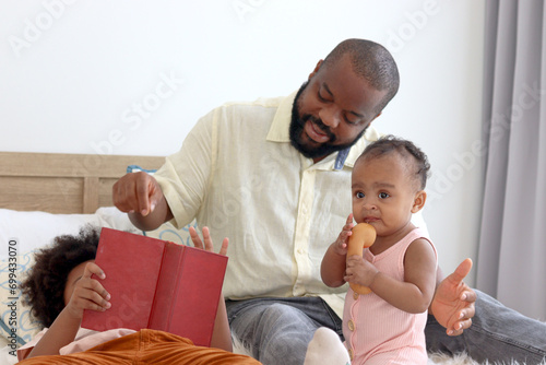 Happy African family, little cute toddle baby infant daughter girl plays with father on white bed while son brother reads in bedroom. Dad looks after child at home, kids and parent spend time together