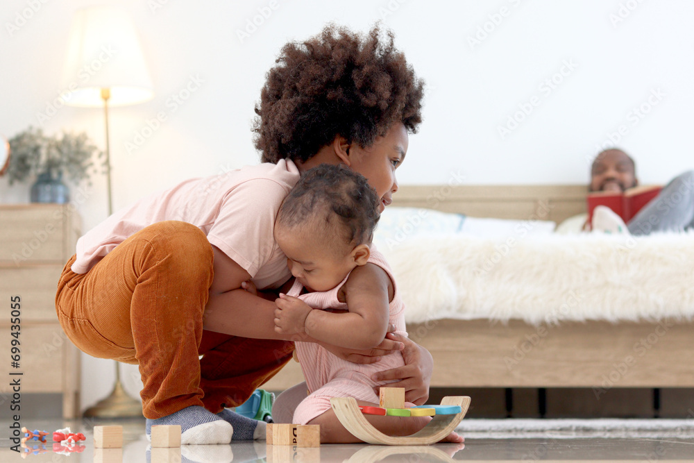 Obraz premium Happy African family spending time together, brother boy with black curly hair try to hold and lift up little cute toddle baby infant kid on floor with blurred background of dad reading book on bed.